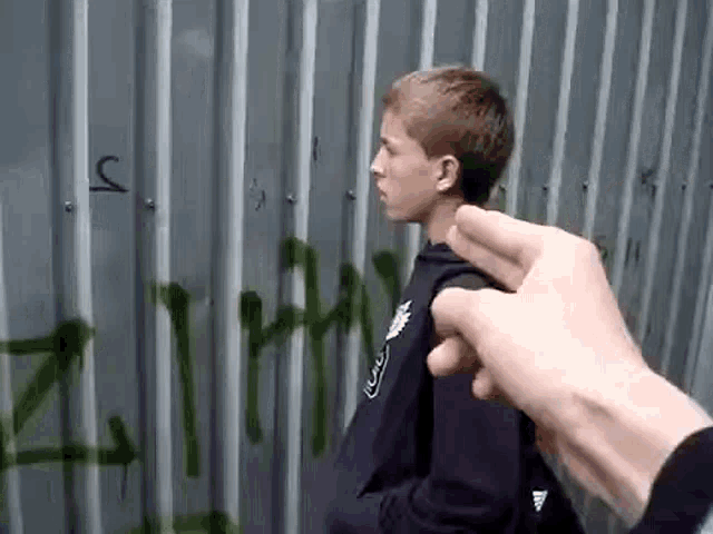 a person is pointing at a young boy standing in front of a fence with graffiti on it .