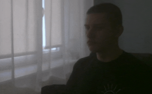 a man in a black shirt is sitting in front of a window with a white curtain .
