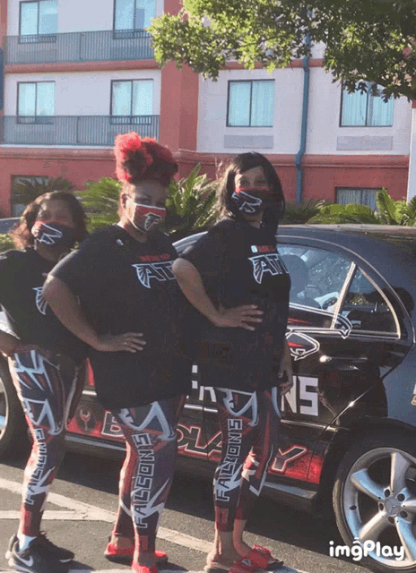 three women wearing face masks stand in front of a car that says falcons on it