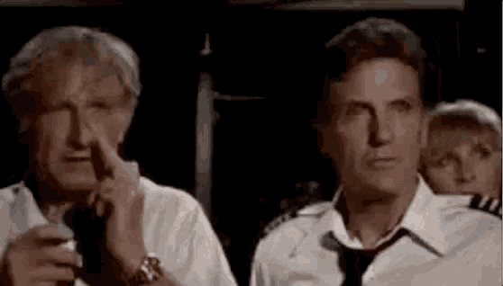 two men are sitting next to each other on an airplane . one of the men is holding his nose .