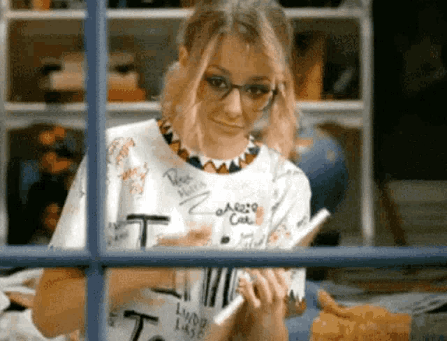 a woman wearing glasses and a shirt with the letter t on it looks out a window