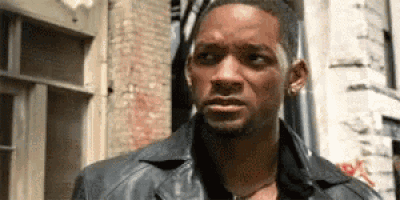 a man in a leather jacket is standing in front of a brick building and looking at the camera .