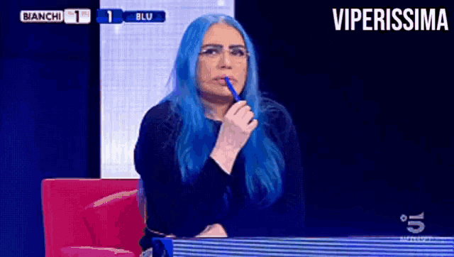 a woman with blue hair holds a pen in her mouth while sitting in front of a television screen that says viperissima
