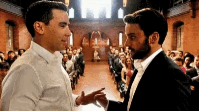 two men in suits are standing next to each other in front of a crowd of people at a wedding ceremony .