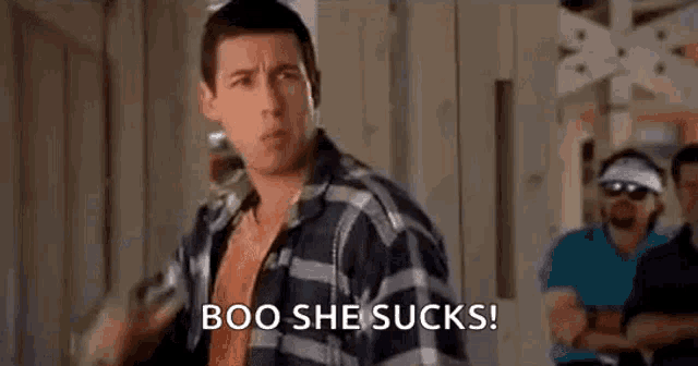 a man in a plaid shirt is saying boo she sucks while standing in front of a group of men .