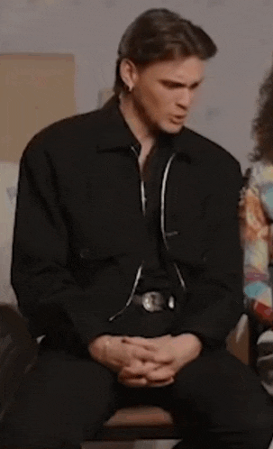 a man in a black jacket is sitting with his hands folded .