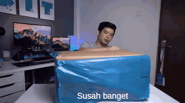 a man sitting at a desk with a box wrapped in blue plastic with the words sudah banget on it