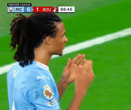 a man with dreadlocks applauds on a soccer field with a scoreboard that says premier league