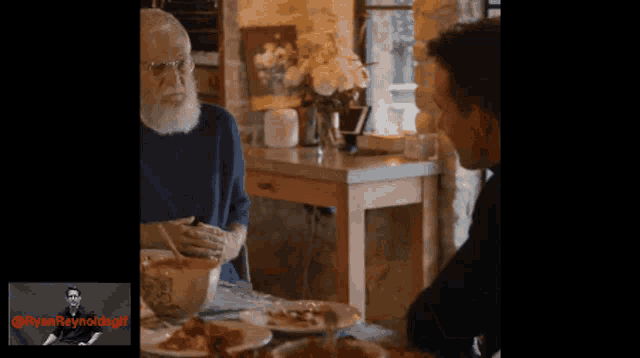a man with a beard sits at a table with plates of food and a picture of ryan reynolds