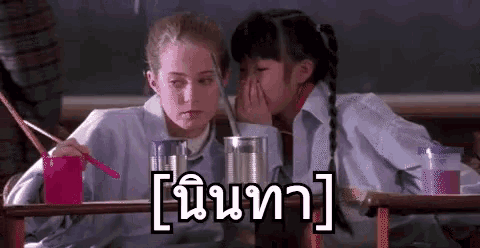 two girls are sitting at a table in a classroom covering their faces with their hands .