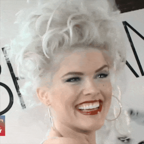 a woman with white hair and red lips smiles in front of a sign that says n