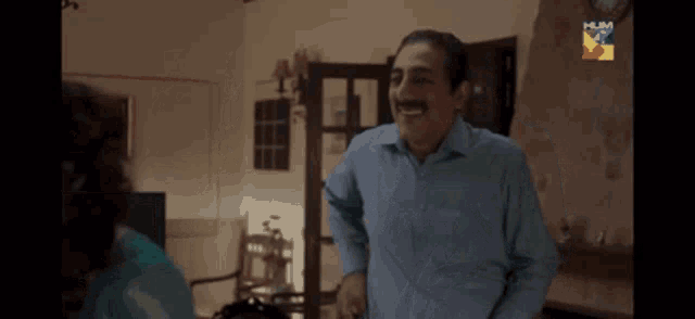 a man in a blue shirt is laughing in a living room with a hum logo behind him