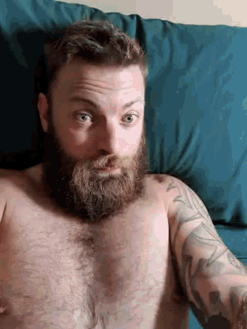 a shirtless man with a beard is laying on a bed with a blue pillow .