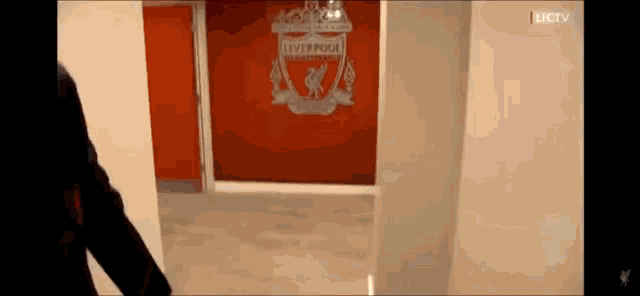 a man is walking through a hallway with a liverpool logo on the wall behind him .
