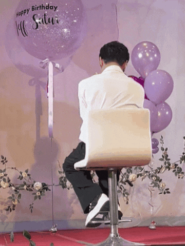 a man sits in a chair in front of purple balloons that say happy birthday saturday
