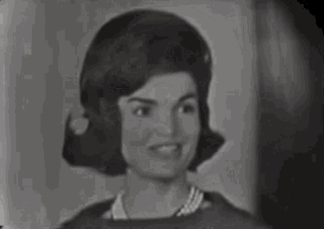 a woman wearing a pearl necklace and earrings is smiling in a black and white photo .