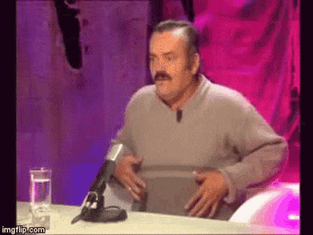 a man with a mustache is sitting at a table with a microphone in front of him .