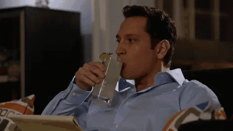 a man in a blue shirt is sitting on a couch drinking a drink from a glass .