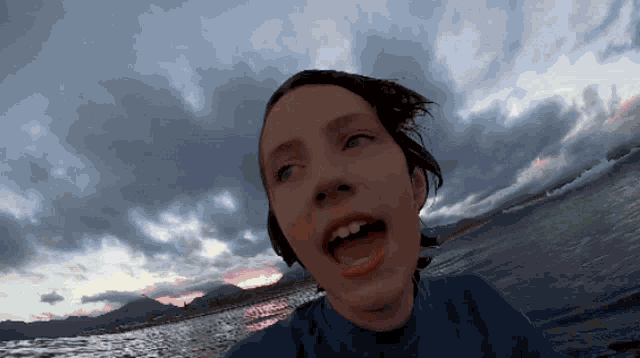 a woman in a blue shirt is standing in front of a body of water with her mouth open