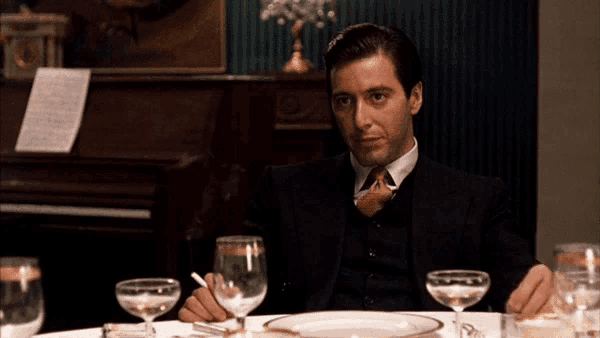 a man in a suit and tie sits at a table with wine glasses and a cigarette