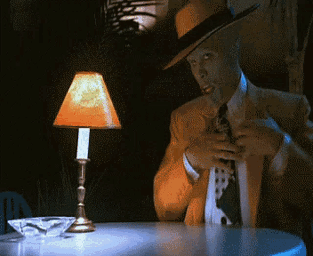 a man in a suit and hat sits at a table with a lamp on it