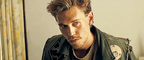 a man with a beard is wearing a green leather jacket and looking at the camera .