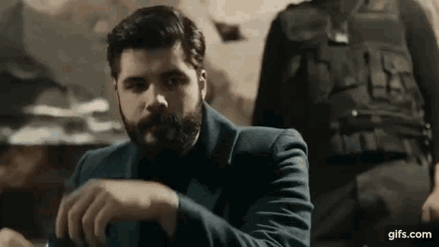 a man with a beard is sitting at a table in a dark room looking at his watch .