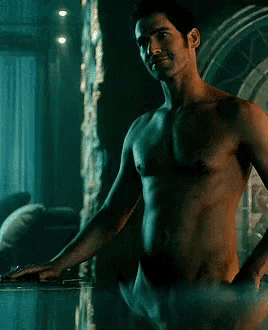 a shirtless man stands in a dark room looking out a window