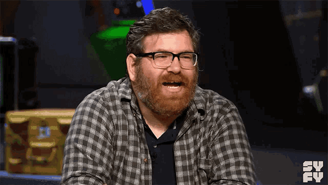 a man with glasses and a beard is wearing a plaid shirt with a tv logo behind him