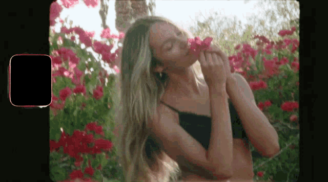 a woman in a black bra is smelling a flower in front of a bush of pink flowers .