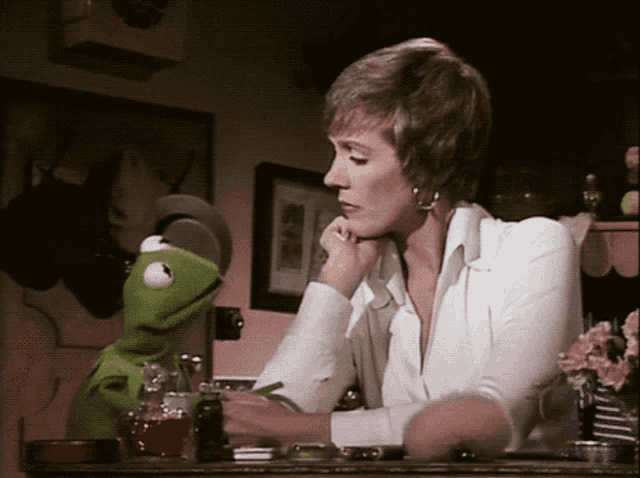 a woman sits at a desk with a kermit puppet