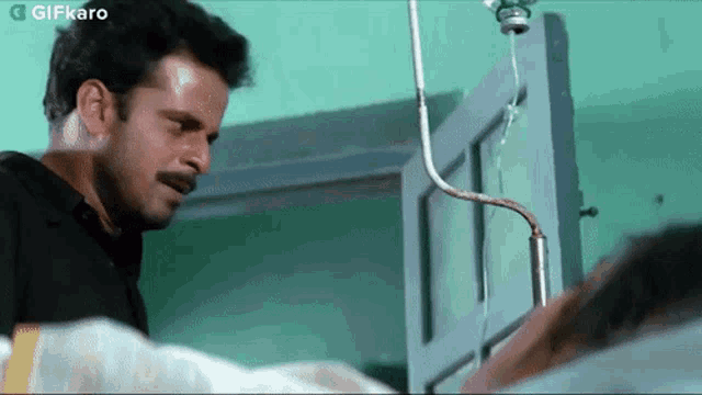 a man with a mustache is standing next to a man in a hospital bed with an iv hanging from the door .