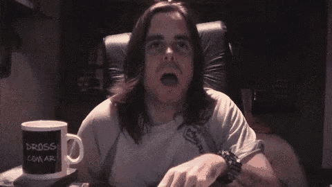 a man with long hair is sitting in a chair with his mouth open and a mug that says " gross " on it