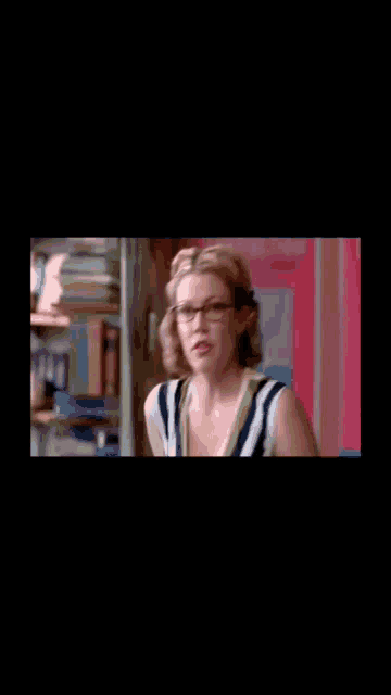 a woman wearing glasses and a striped shirt is standing in front of a bookcase .