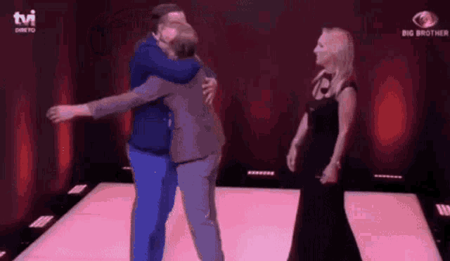 a man in a suit is hugging another man while a woman in a black dress stands behind them ..