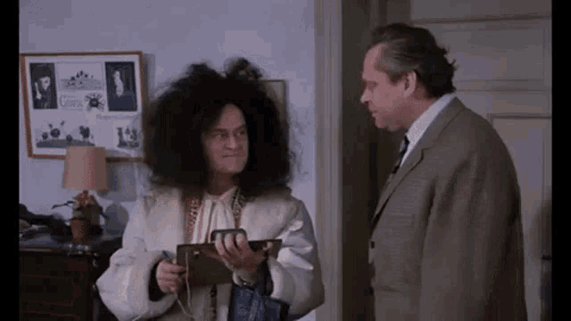 two men are standing next to each other in a room . one of the men is wearing a wig .