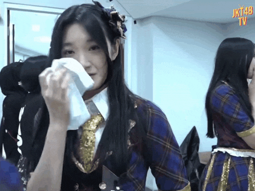 a girl wipes her face with a napkin in front of a jkt48 tv screen