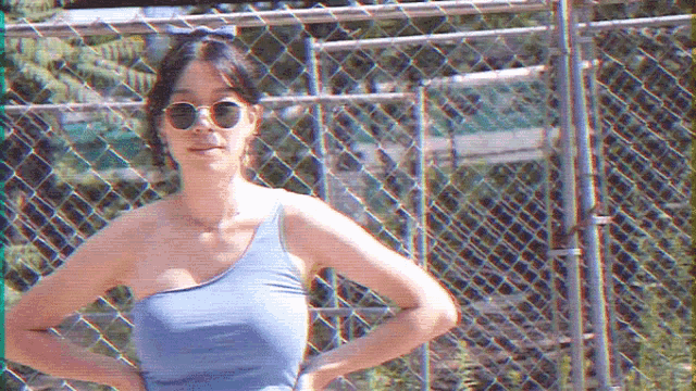 a woman wearing sunglasses and a blue one shoulder top is standing in front of a chain link fence