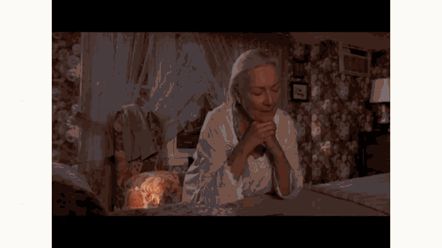 an older woman is sitting on a bed with her hands folded in prayer .