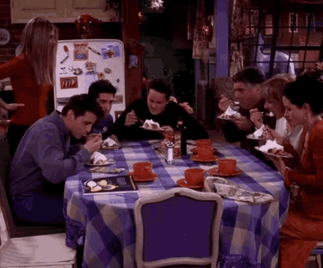 a group of people are sitting at a table with plates of food and a refrigerator with a picture of friends on it