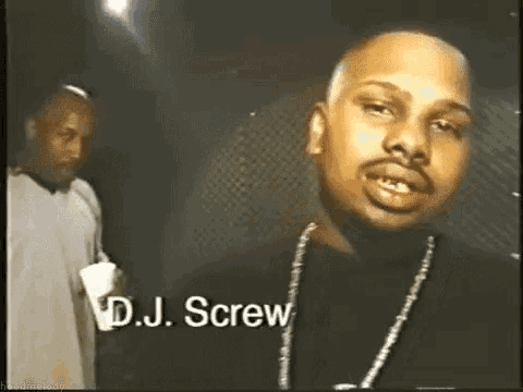 a man wearing a necklace with the name dj screw on it is standing next to another man in a dark room .