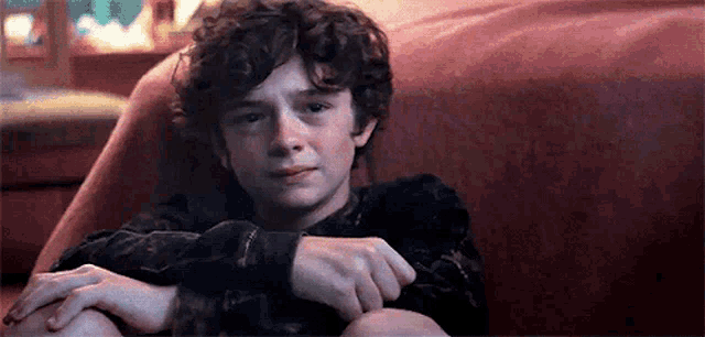 a young boy with curly hair is sitting on a red couch with his hands on his knees .