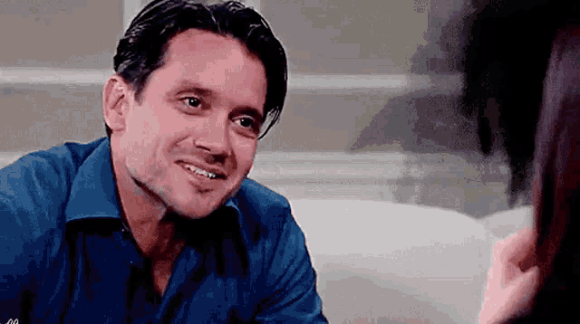 a man in a blue shirt is smiling while sitting on a couch talking to a woman .