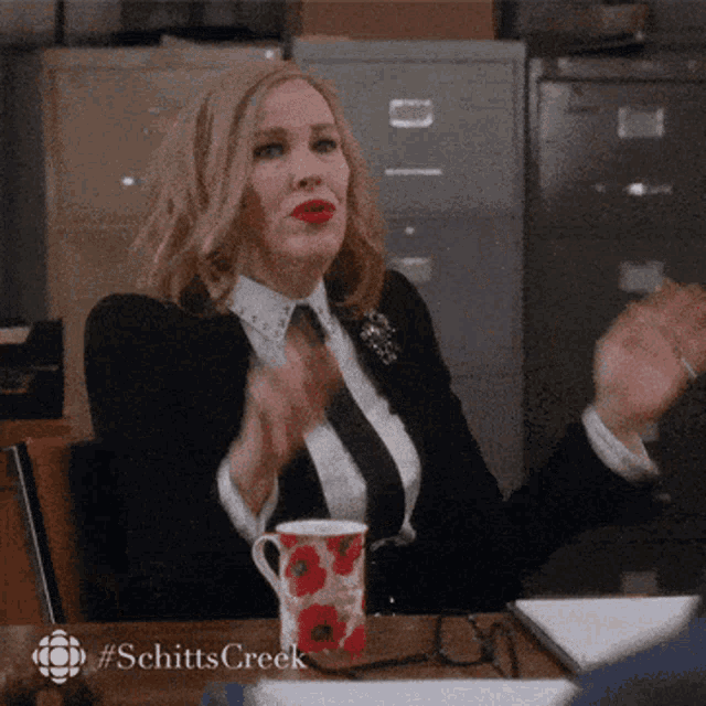 a woman in a suit and tie is sitting at a desk with a cup of coffee and a schitts creek logo