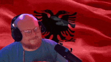 a bald man wearing headphones and glasses stands in front of a red flag