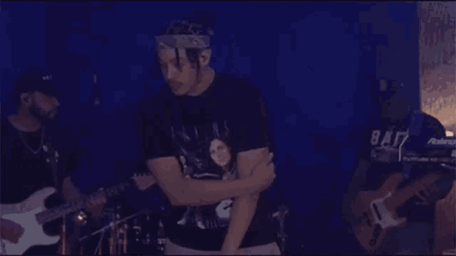a man wearing a bandana is standing in front of a man playing a guitar and another man playing drums .