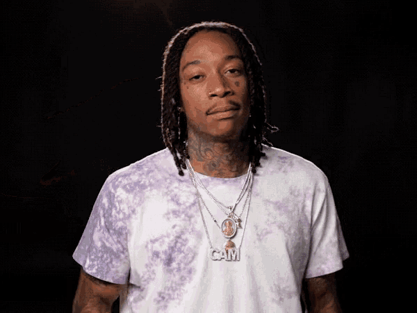 a man wearing a purple tie dye shirt has a necklace with the word cam on it