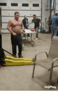 a man without a shirt is standing next to a chair and a table in a garage .