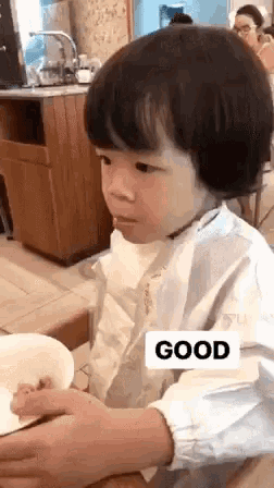 a little boy is sitting at a table with a bowl of food and a sign that says good on it .
