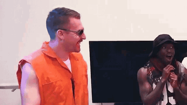 two men are standing next to each other in front of a television . one of the men is wearing an orange vest .